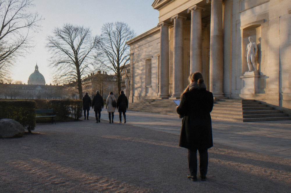 A guide collecting tips from departing walkers beside a historic Copenhagen building at the end of a tour