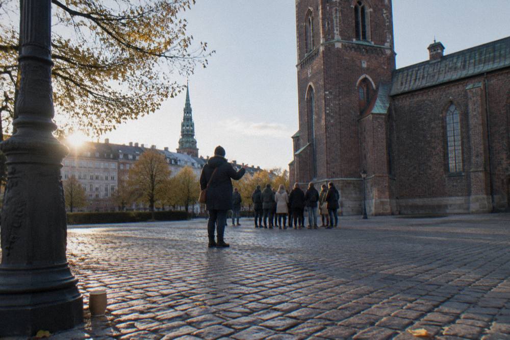 A guide speaking with a small group of visitors on a cobblestone square beside an old Copenhagen church tower