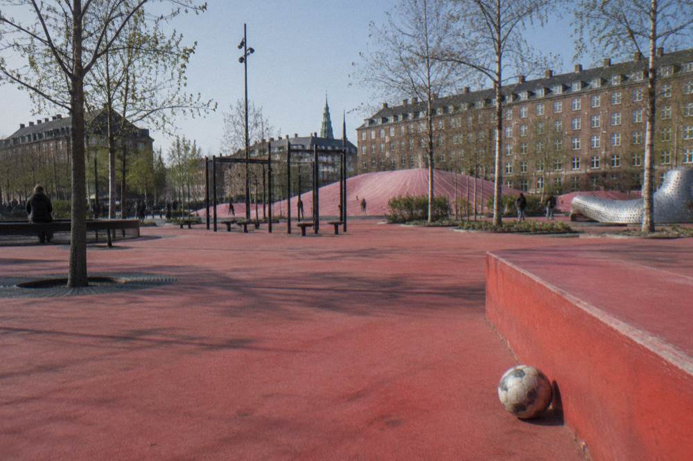 Un parque urbano amplio con suelo rojo brillante, bancos y equipos de juego modernos bajo el cielo abierto