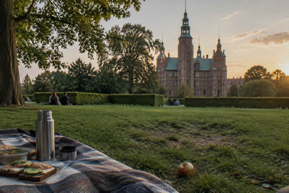 A picnic blanket on a grassy park lawn with a packed lunch of rye bread sandwiches and a thermos beside old castle walls