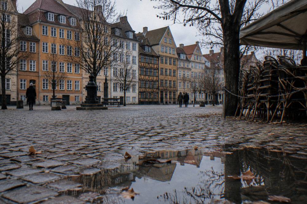 Cobbled square of Gråbrødretorv on a flat grey winter afternoon, ochre and cream townhouses, bare lime trees, stacked cafe chairs and a puddle reflecting the gables