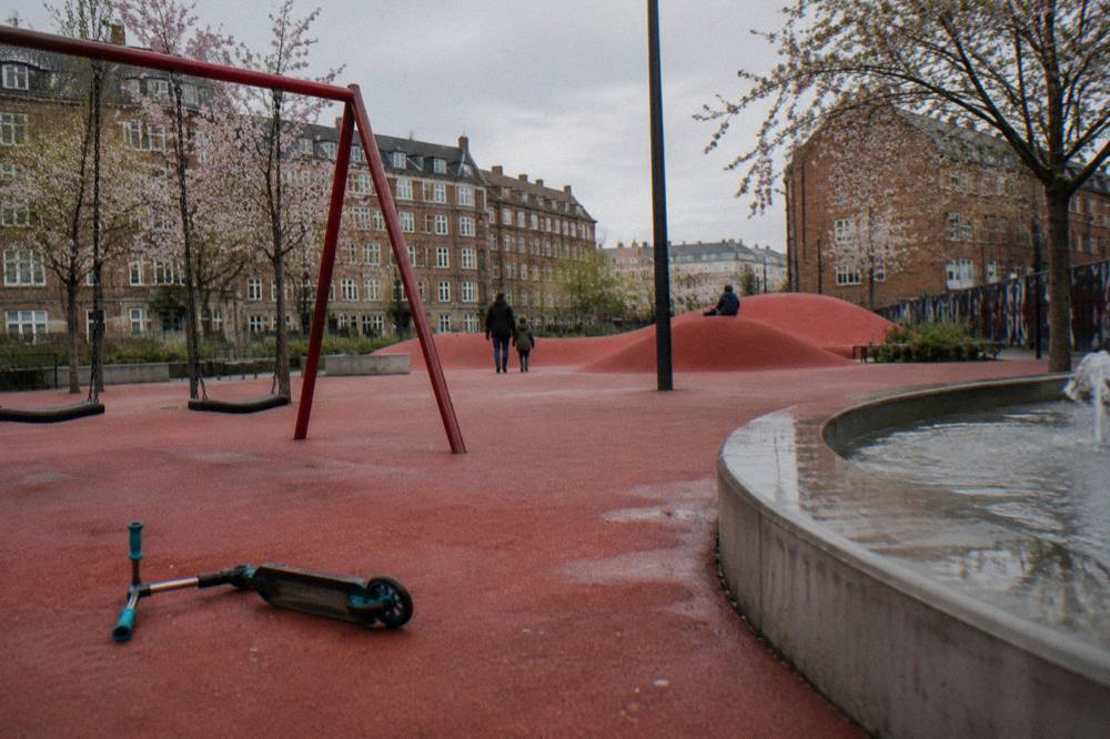 Bright red paved square of a public park with benches, swings and a fountain